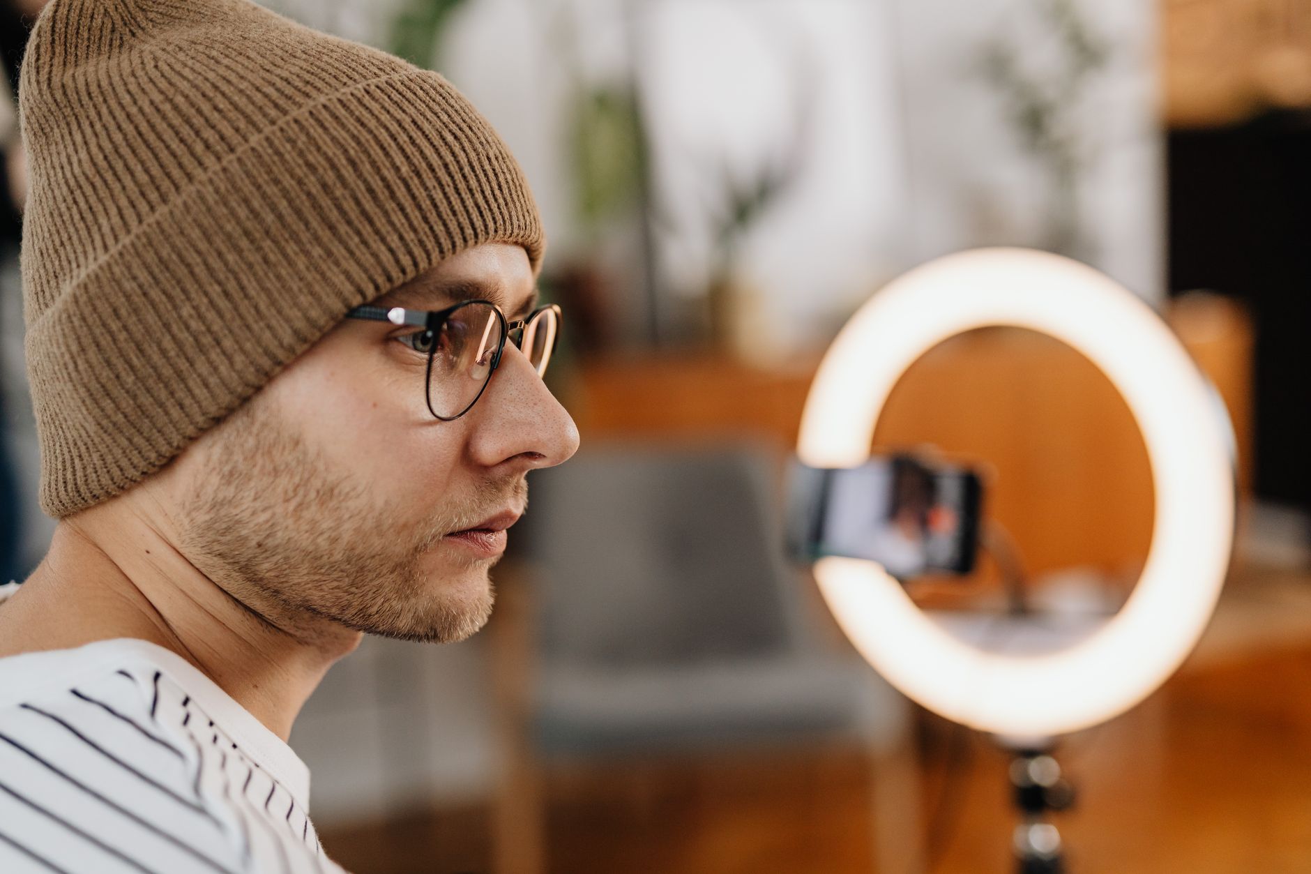 Un homme à lunettes et bonnet marron, de profil, face à une lampe annulaire équipée d’un smartphone.