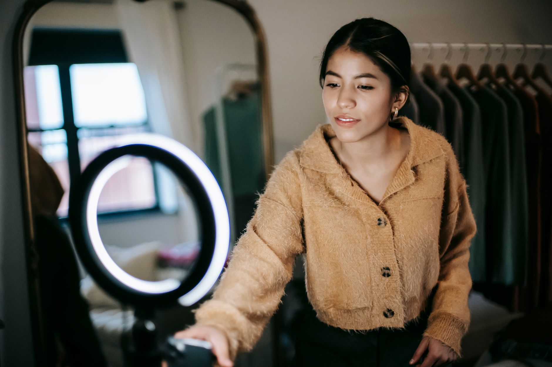 Une jeune femme en gilet beige sourit près d’une lampe annulaire, avec un grand miroir et un portant de vêtements derrière elle.