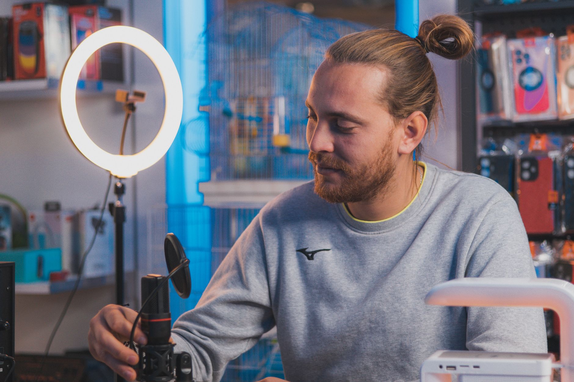 Un homme aux cheveux attachés en chignon ajuste un micro devant une lampe annulaire allumée.