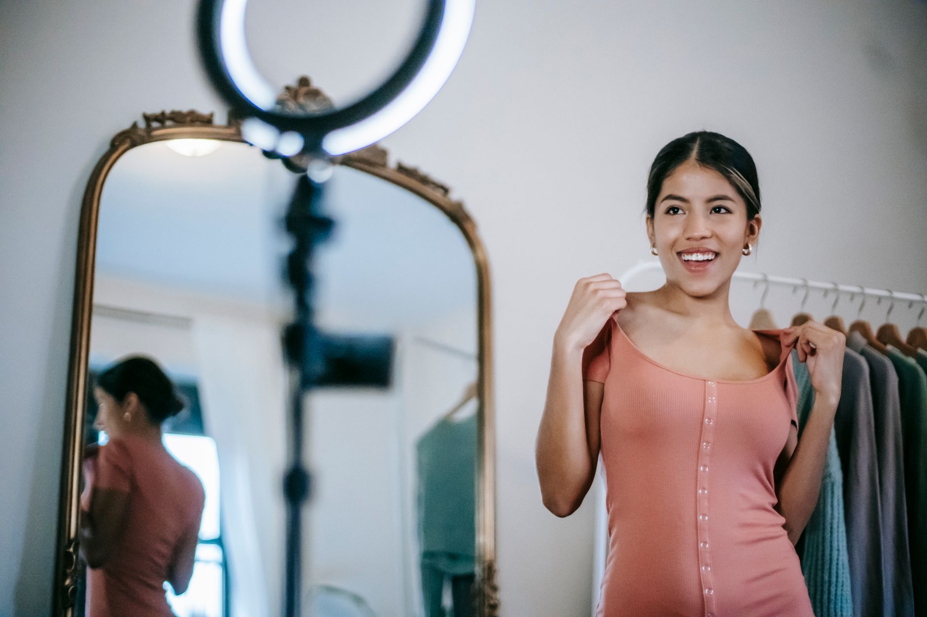 Une jeune femme souriante en haut rose se filme devant un grand miroir doré, une lampe annulaire au-dessus d’elle.