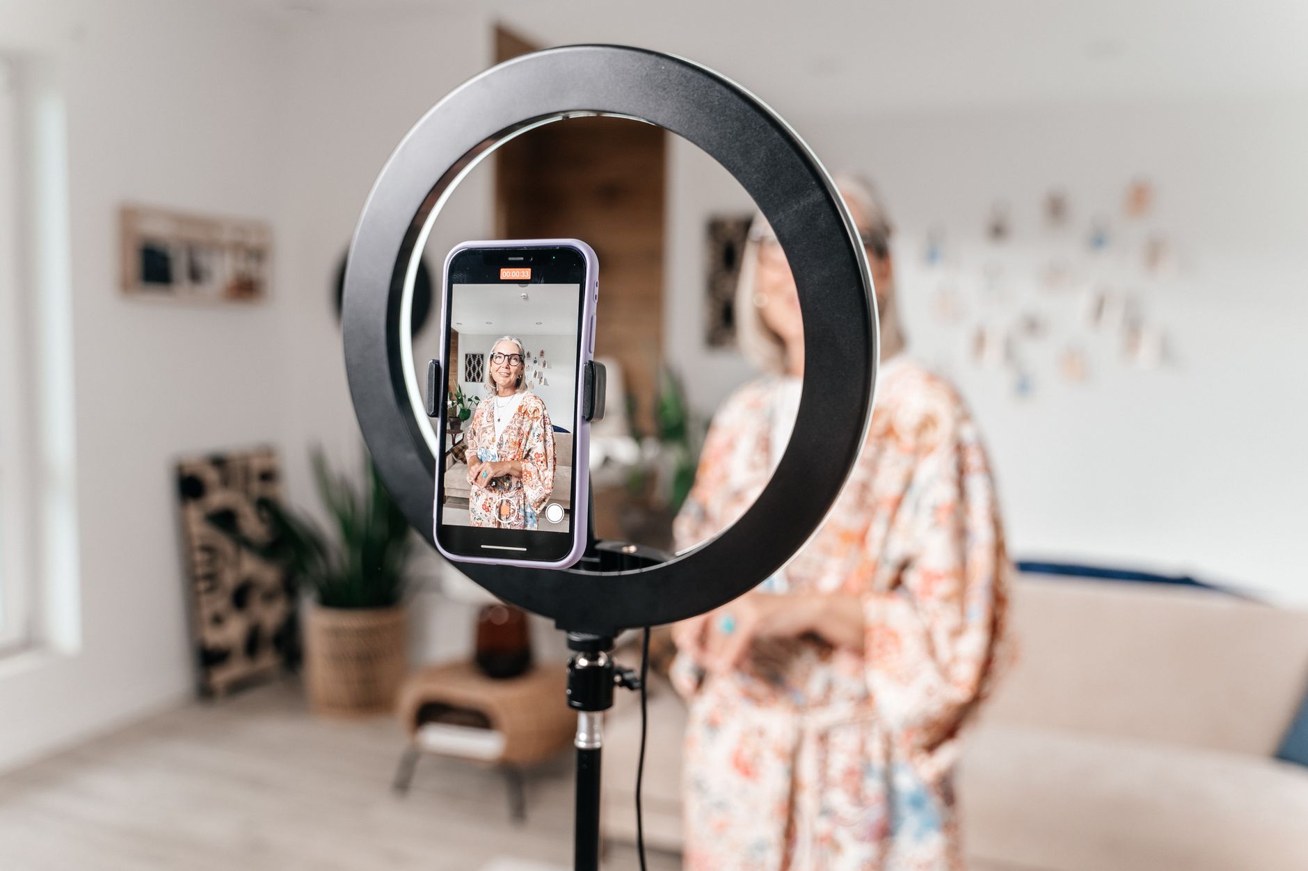 Un smartphone fixé sur une lampe annulaire filme une femme aux cheveux gris dans un salon lumineux.